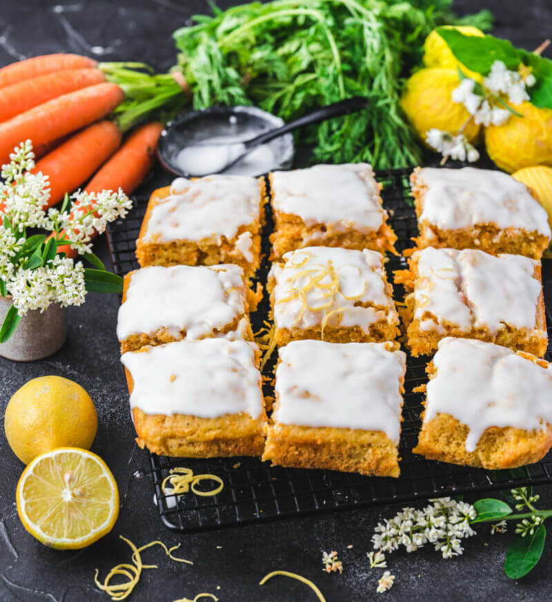 Veganer Karottenkuchen in Blechform, geschnitten in neun gleich große Stücke und garniert mit Zuckerguss, angerichtet auf einem Blech und dunklem Hintergrund