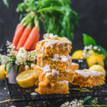 Mehrere Stücke des veganen Karottenkuchens aufeinandergestapelt und mit Zuckerguss garniert, angerichtet auf einer dunklen Marmorplatte und dunklem Hintergrund