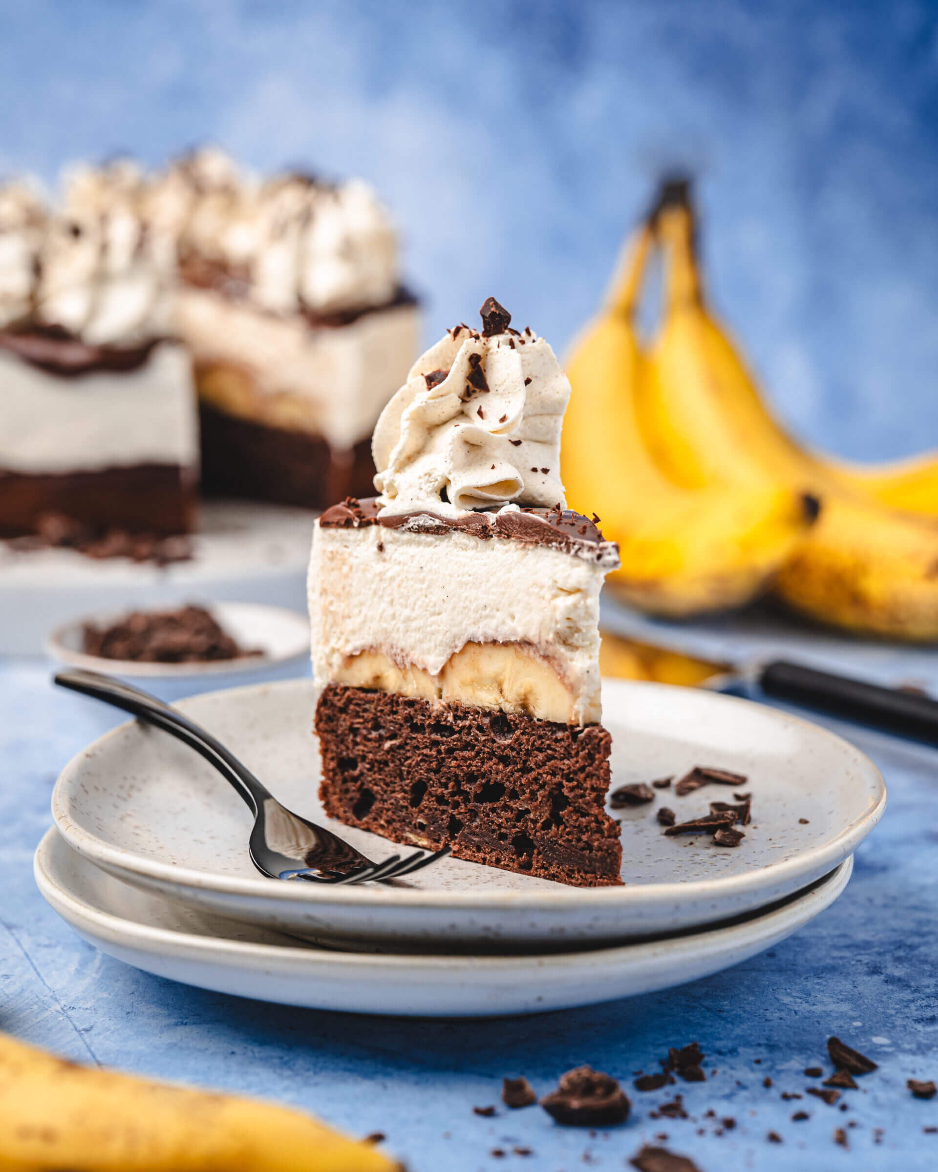 Ein Stück Schokoladen Bananen Torte auf blauen Untergrund, im Hintergrund die Torte mit dem fehlenden Stück und ein Bund Bananen